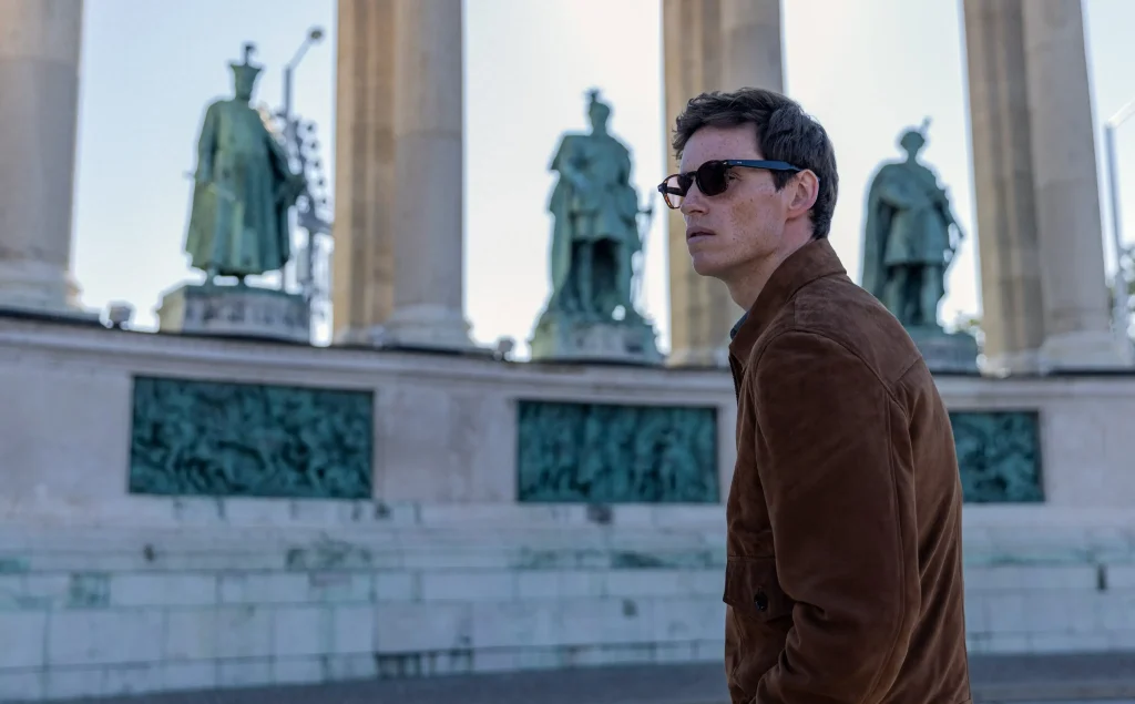 Depiction from "The Day of the Jackal" series with a man in a brown jacket and sunglasses, suggesting intrigue and suspense.