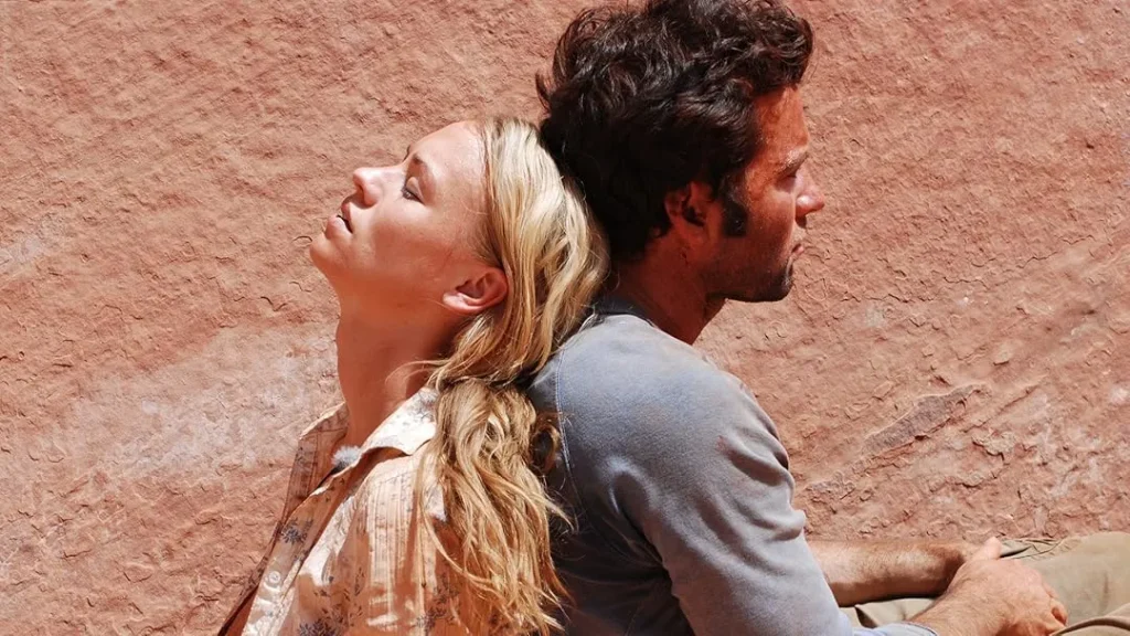A man and woman sit back to back on a rocky ledge, surrounded by the vast expanse of a canyon landscape.