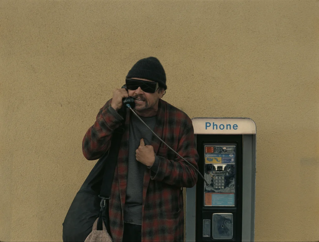An angry man next to a payphone, engaged in a phone call, reflecting frustration in a war epic context.