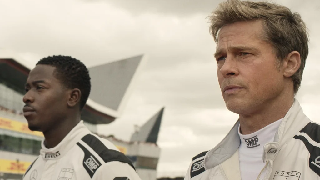 Brad Pitt and his teammate in cool race outfits, leaning against a high-performance F1 car from Drive to Survive.