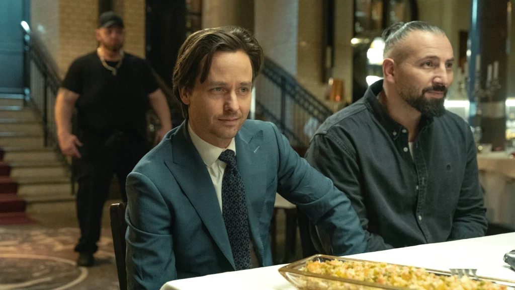 A scene from "Murder Mindfully" shows a man about to eat, while a severe-looking man sits nearby.