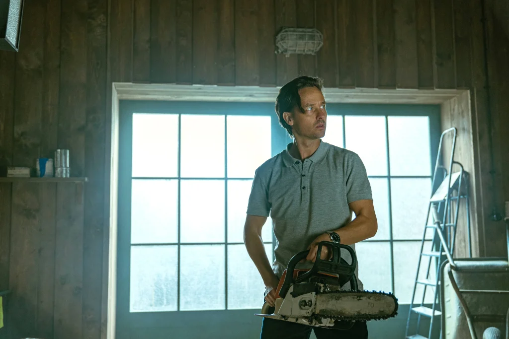 A man stands in front of a window, holding a chainsaw, appearing ready to act after a murder in the series "Murder Mindfully."